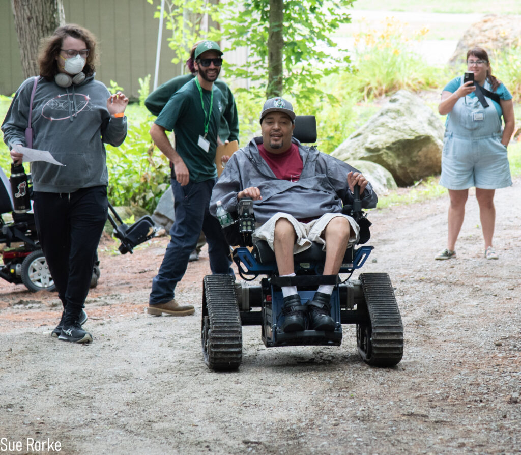 Man in trail wheelchair starts down trail with friend. DCR staff look on