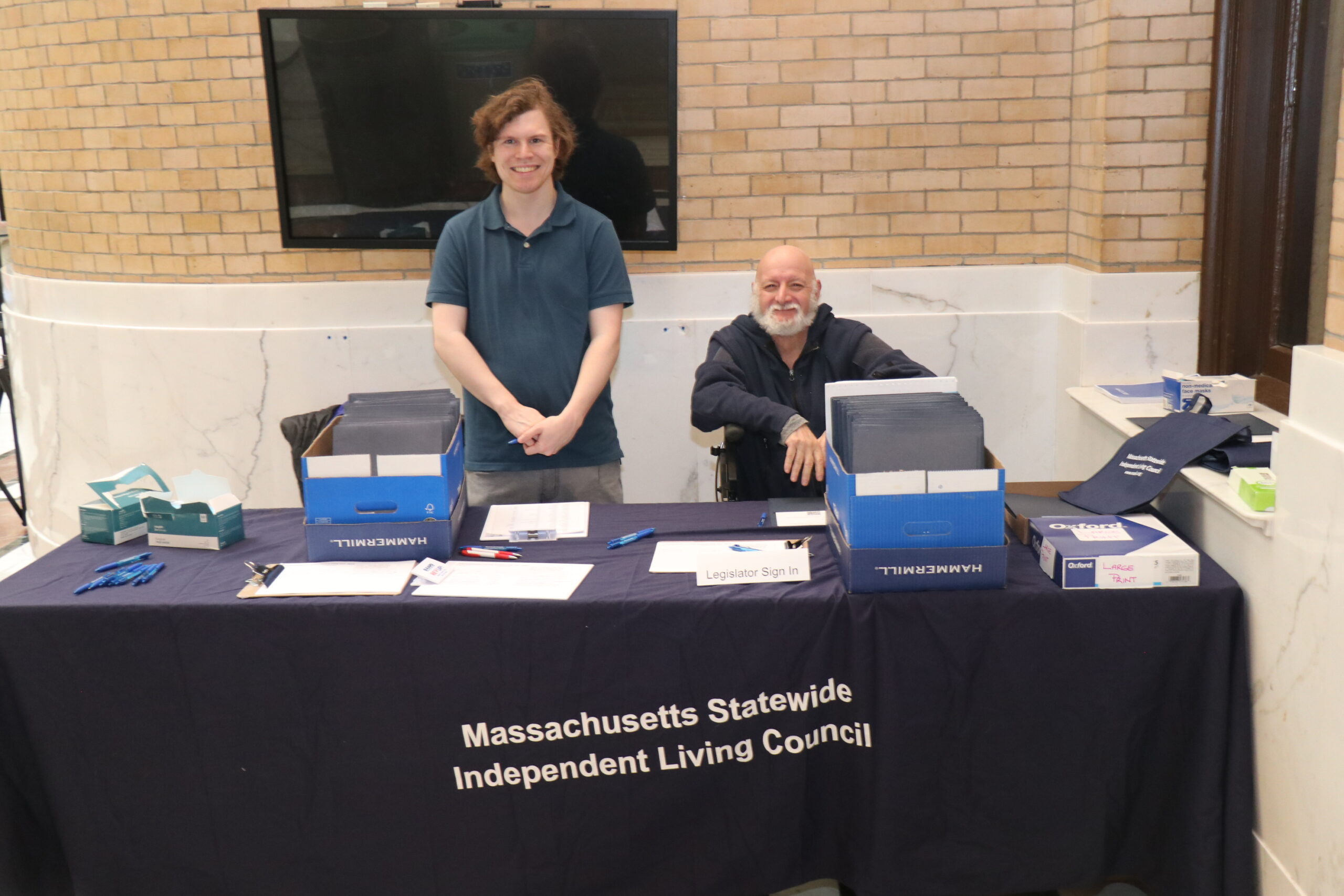 Ian Hacker and Jim Tozza smiling from behind the sign in table, which has a tablecloth reading "Massachusetts Statewide Independent Living Council" , and folders, papers and pens on top