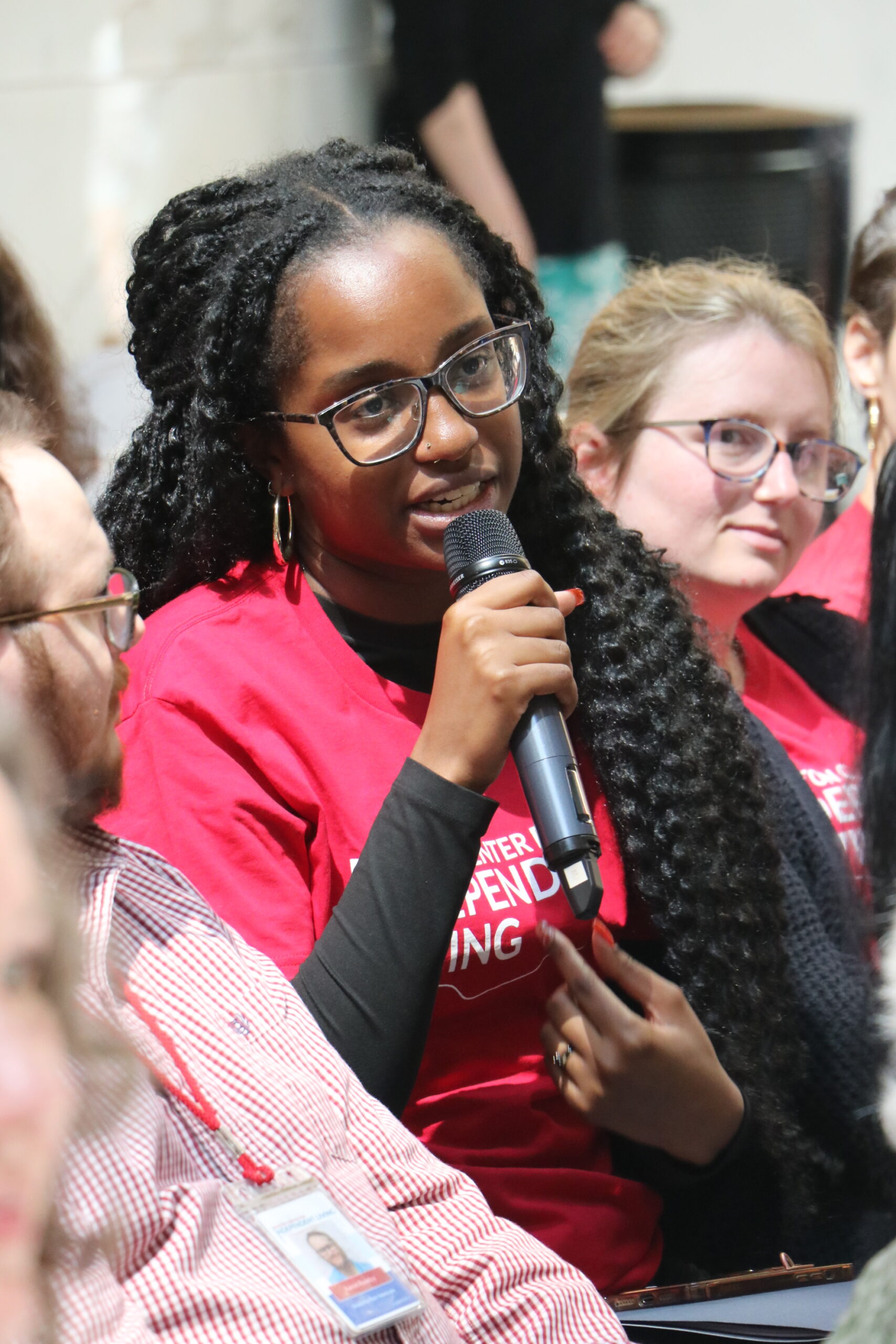 Someone wearing a Boston Center for Independent Living t-shirt speaking into a microphone, seated next to other people in the audience