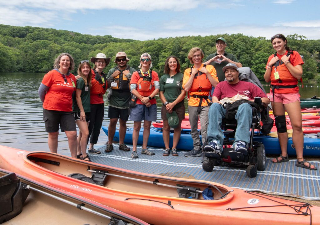 A group of people smiling in front of the lake, some wearing personal floatation devices (aka life jackets)
