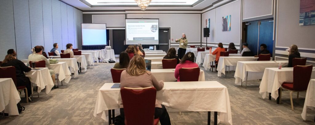 A workshop room, with people sitting at tables and chairs facing a presentation at the front of the room