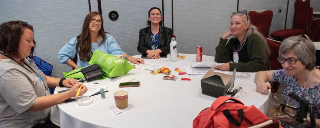 A group of people sitting around a table, smiling, one writing
