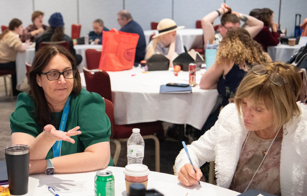 Two people at a table, one gesturing, the other writing